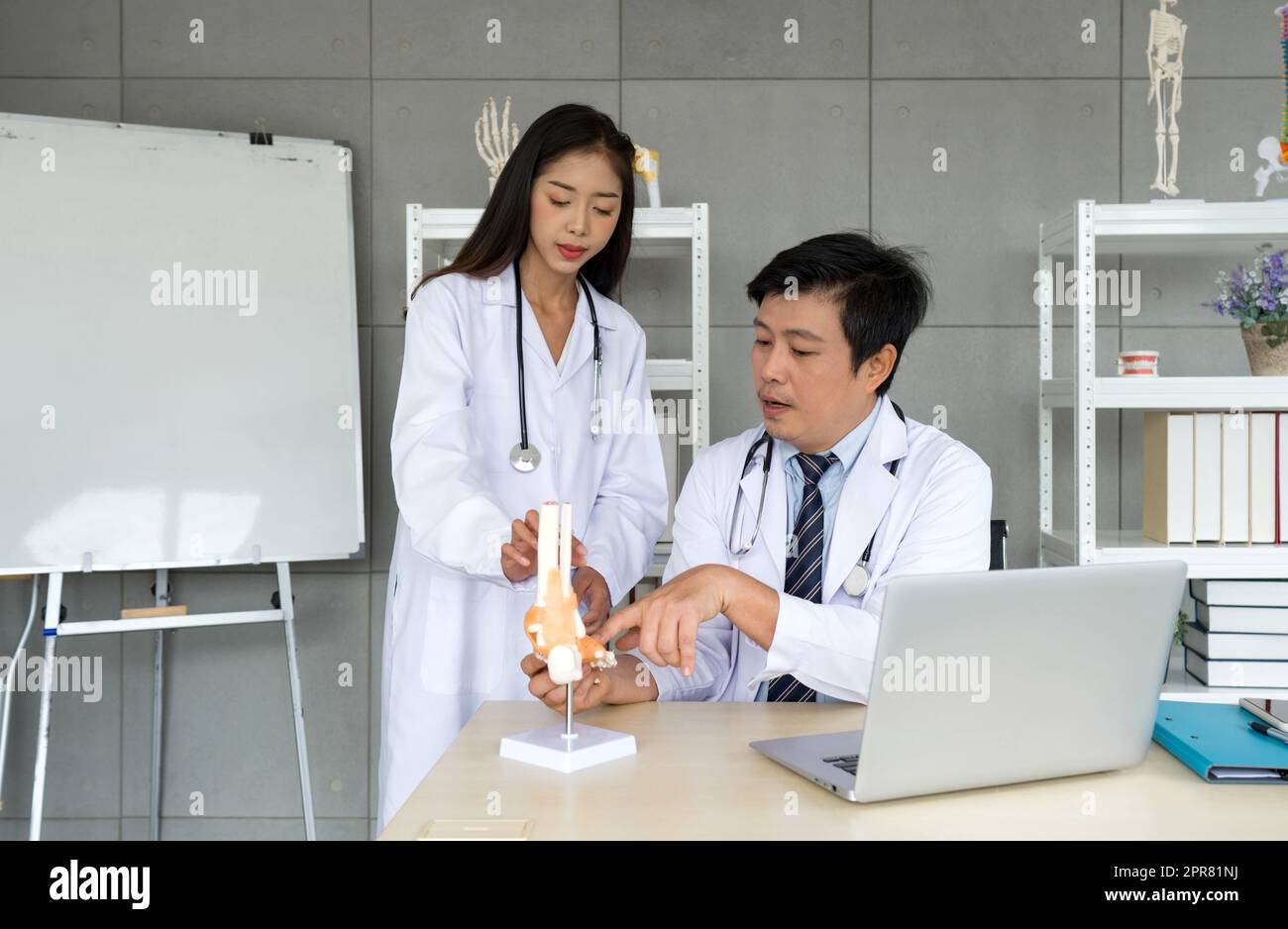 Giovani chirurghi ortopedici asiatici in abito bianco e stetoscopio che chiedono al professore consigli su ossa, articolazioni, tendini e muscoli. Atmosfera di lavoro in ospedale. Foto Stock