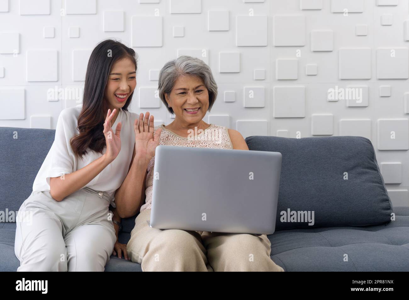 La giovane donna asiatica e la nonna agitavano la mano attraverso la comunicazione remota attraverso il computer portatile nel soggiorno. Felice famiglia che vive insieme Foto Stock