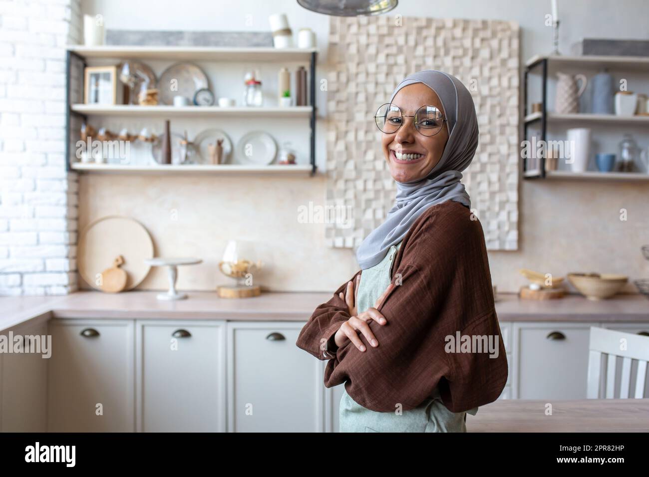 Ritratto di una giovane casalinga araba in hijab in piedi in cucina in grembiule, braccia incrociate sul petto, sorridendo alla macchina fotografica. Foto Stock