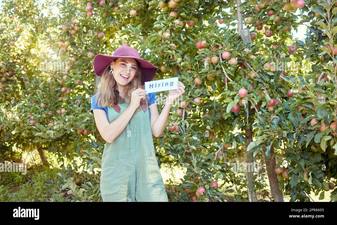 Contadina che reclutava operaie per aiutare la sua startup. Giovane donna felice con un cartello di assunzione in una fattoria vicino ai frutteti di mele. Carenza di manodopera agricola, mercato del lavoro agricolo e occupazione Foto Stock