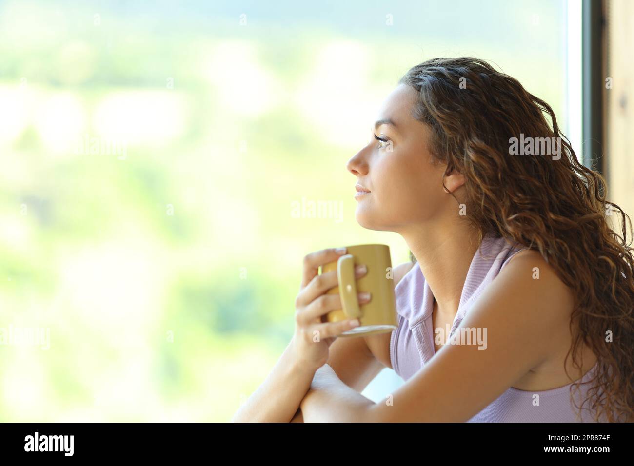 Donna che tiene una tazza di caffè che si rilassa guardando da una finestra Foto Stock