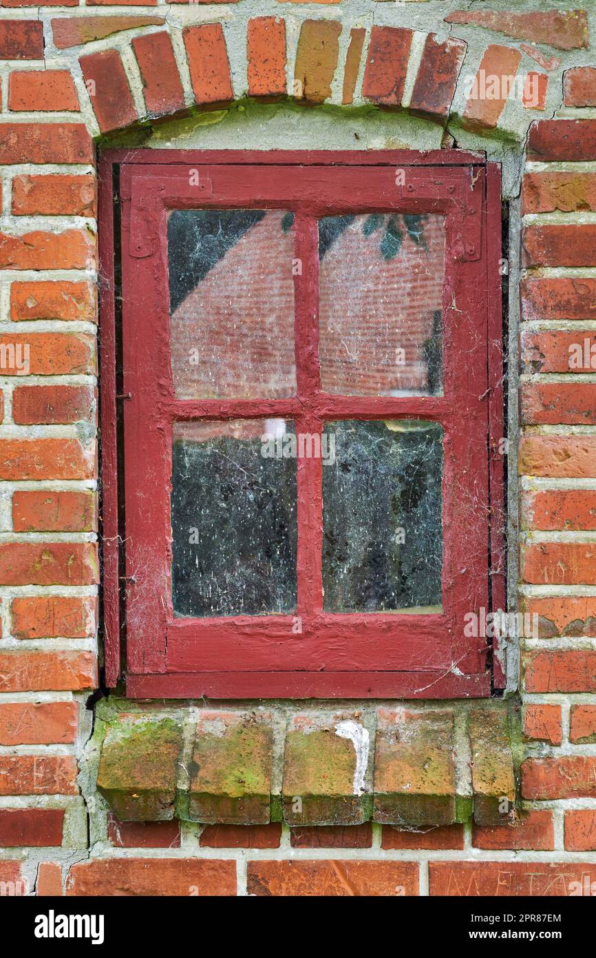 Primo piano della finestra rossa abbandonata coperta da ragnatele, alghe e muschio da abbandono, povertà e crisi economica. Vecchio edificio residenziale vuoto o casa in un villaggio con muffa umida sul davanzale Foto Stock