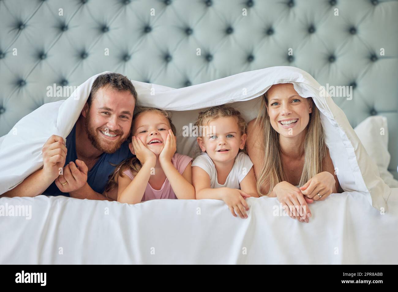 Una famiglia felice a letto sotto una coperta a casa. Ritratto di giovani genitori sorridenti che si divertono con i bambini in camera da letto, coperto da un piumone. Bambine carine che giocano con la madre e il padre Foto Stock