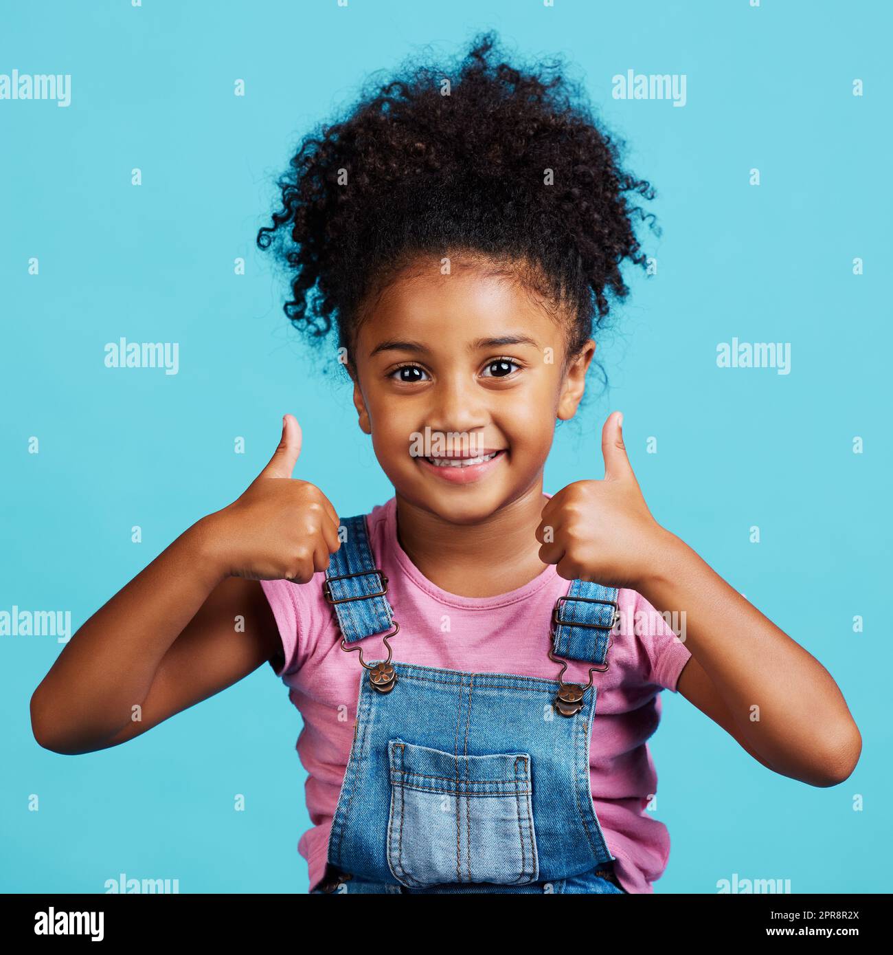 Siate felici, una bambina adorabile che mostra i pollici in piedi contro uno sfondo blu. Foto Stock