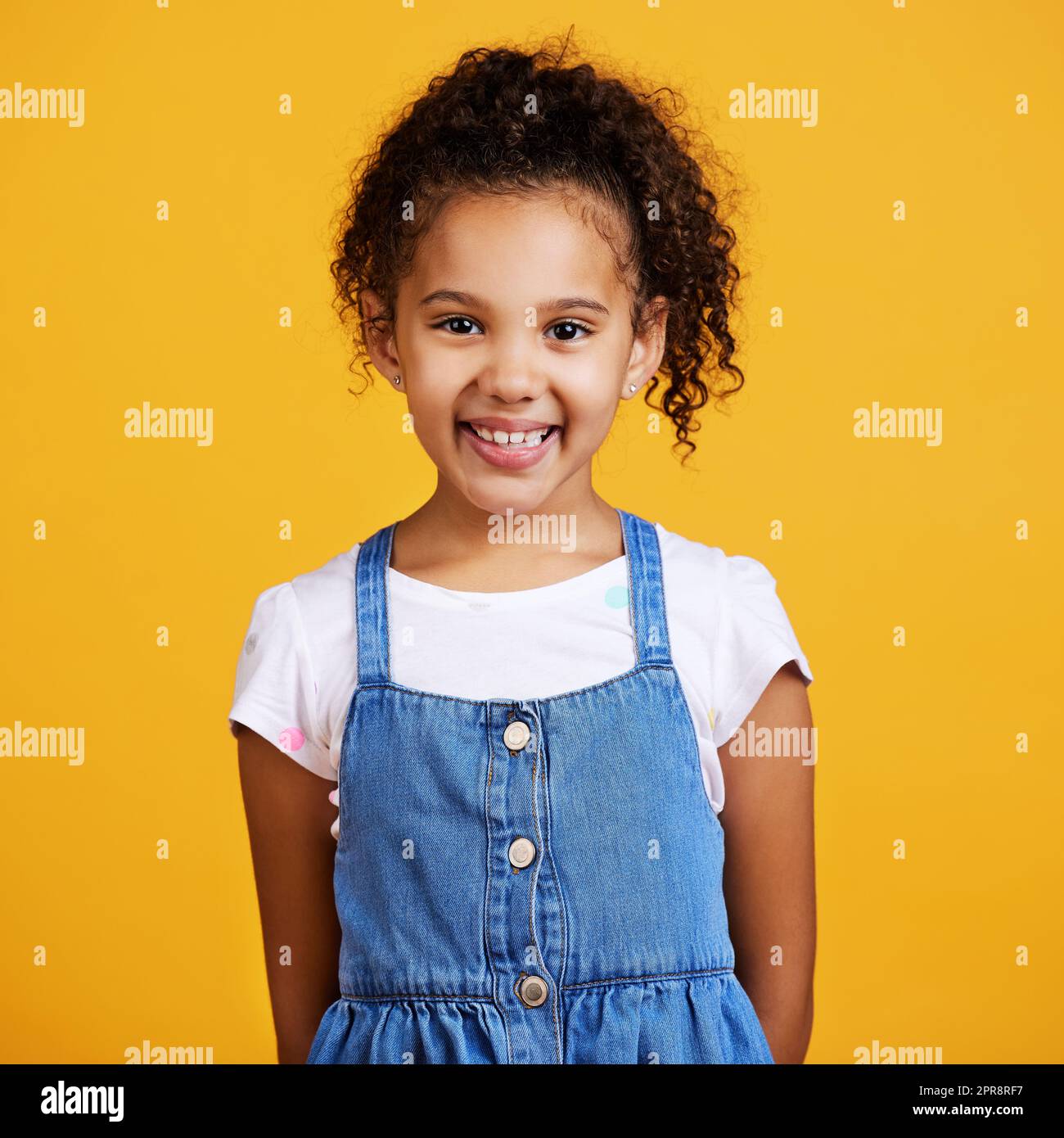 Ritratto in studio di una ragazza di razza mista che sembra in piedi da sola isolata su uno sfondo giallo. Carino bambino ispanico che si posa all'interno. Bambino felice e carino sorridente e spensierato in abiti casual Foto Stock