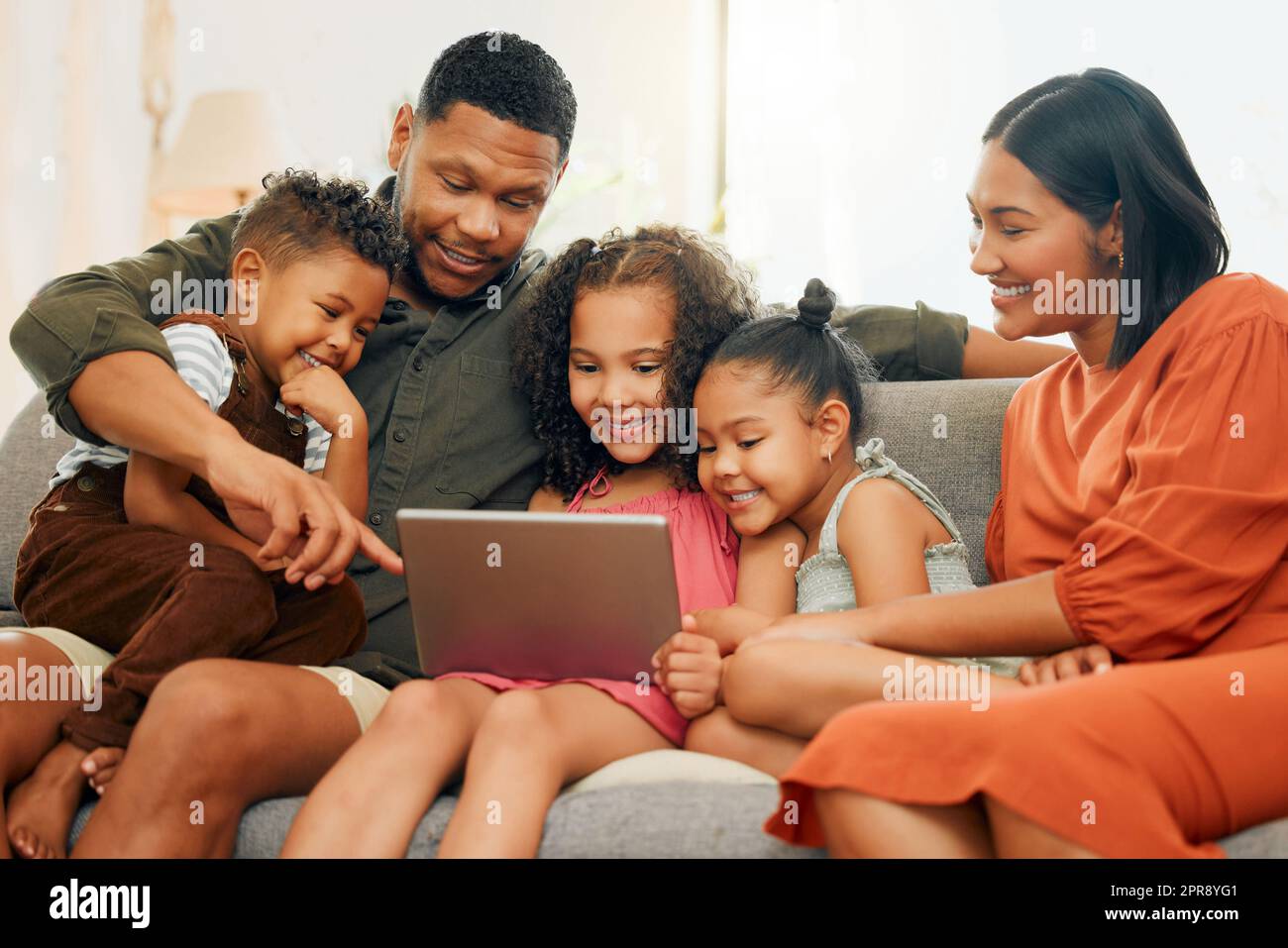 Una felice famiglia di cinque persone che si rilassa sul divano a casa. Ama la famiglia nera essere affezionata sul divano mentre utilizza un tablet digitale e lo streaming. Giovani coppie che legano con i loro figli e guardano film a casa Foto Stock