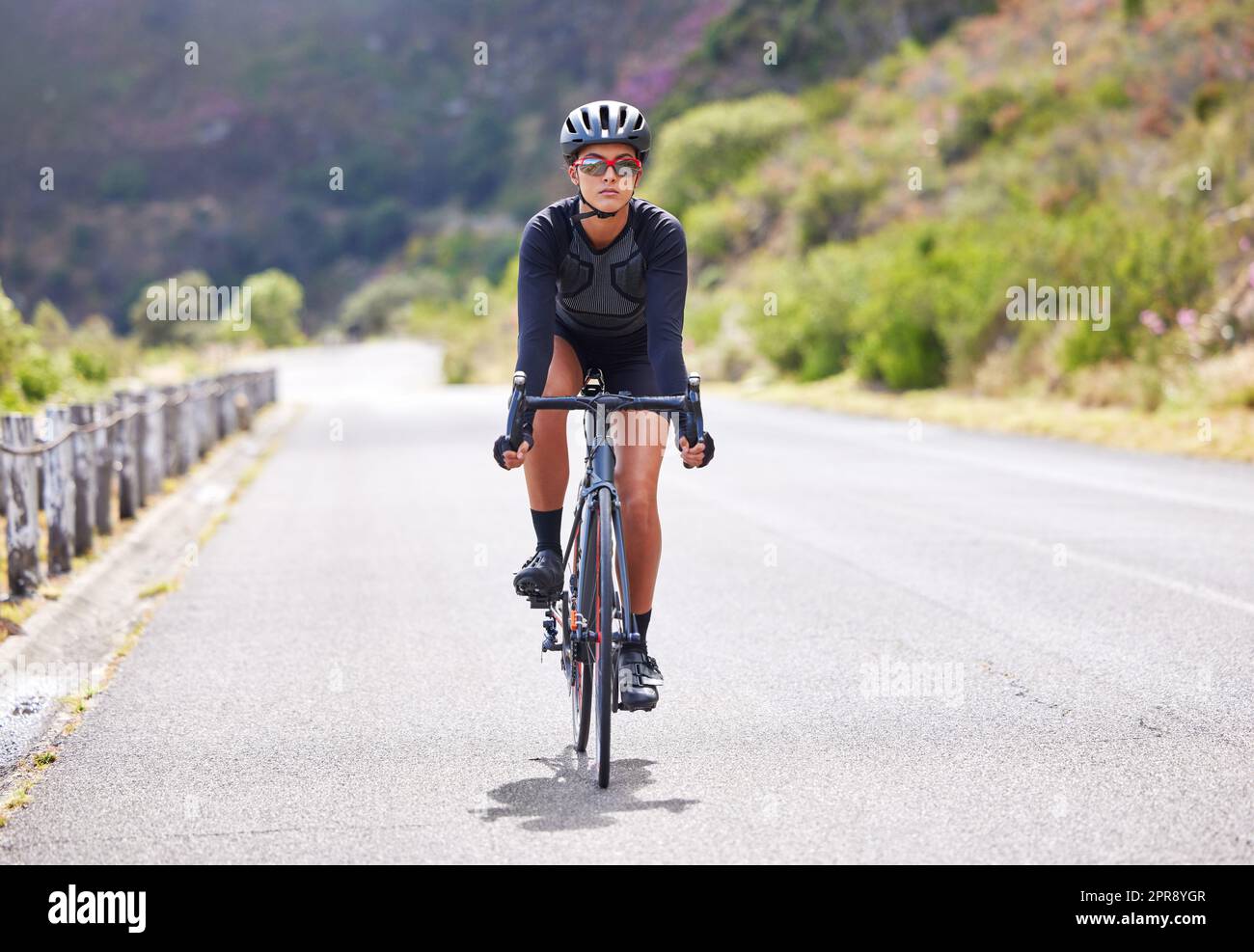 Una giovane donna atletica che va in bicicletta all'aperto. Calzata sportiva femminile che indossa casco e occhiali mentre si guida una bicicletta su una strada lungo la montagna per esercitarsi. Resistenza e cardio durante un allenamento e un allenamento Foto Stock