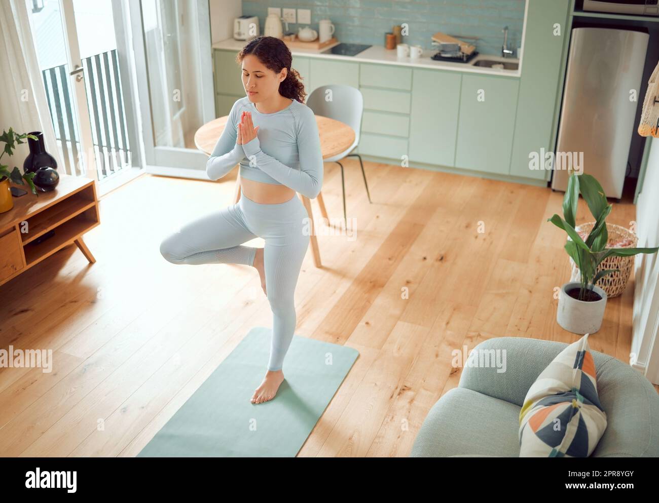 Bella giovane donna mista che medita mentre si trova in posizione eretta mentre pratica yoga a casa. Donna ispanica che esercita il suo corpo e la sua mente, trovando pace interiore, equilibrio e chiarezza Foto Stock