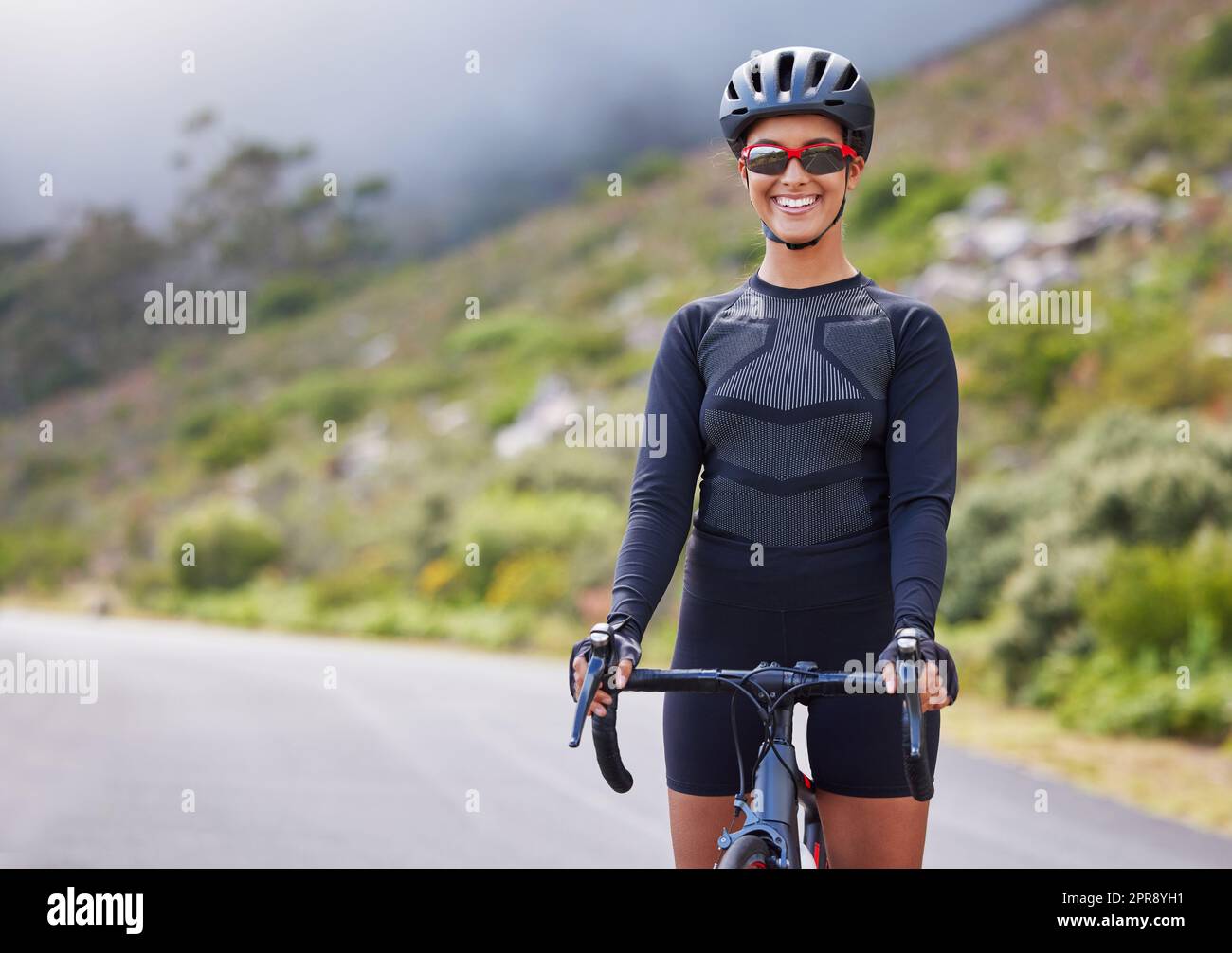 Una giovane donna atletica che va in bicicletta all'aperto. Donna sportiva che indossa un casco e occhiali mentre si guida una bicicletta lungo la montagna per fare esercizio fisico. Sorridente e felice di godersi un hobby Foto Stock
