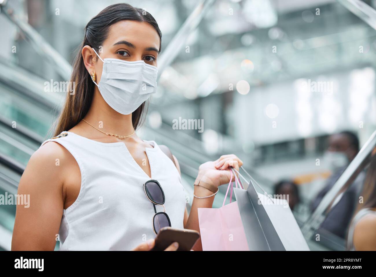 Ritratto di una giovane donna di razza mista che indossa una maschera medica per la prevenzione contro il coronavirus mentre tiene un cellulare e borse della spesa. Alla moda ispanica che trasporta borse al dettaglio dopo l'acquisto in un centro commerciale durante la pandemia di Covid-19 Foto Stock
