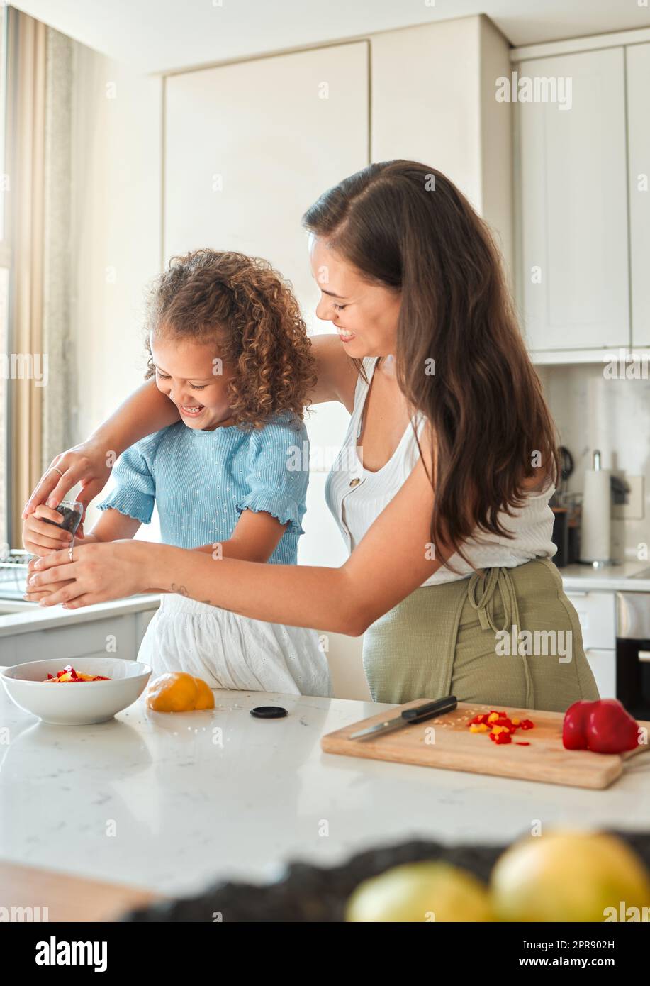 Felice madre insegnando a piccola figlia di cucinare in cucina a casa. Bambina che aggiunge il macinacaffè di pepe condimento in una ciotola mentre fa un'insalata con la mamma Foto Stock