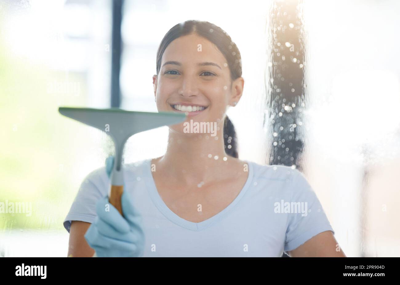 Ritratto di un felice lavoratore domestico misto corsa utilizzando un cigolone su una finestra. Una donna ispanica che ama fare le faccende nel suo appartamento Foto Stock