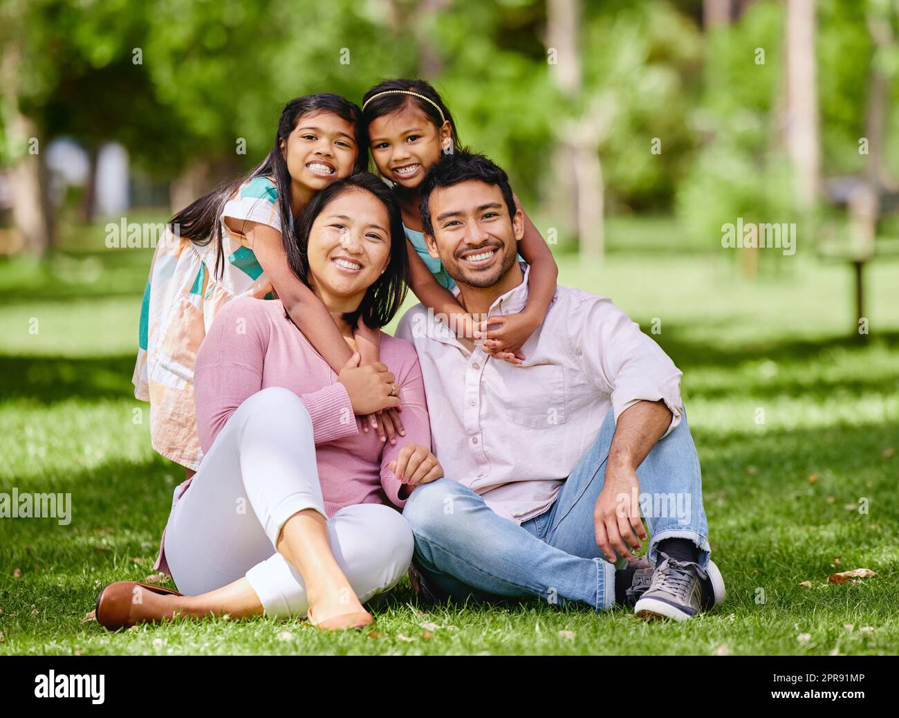 Ritratto della felice famiglia asiatica nel parco. Adorabili bambine che legano e abbracciano i loro genitori fuori in un parco. Marito e moglie a figura intera seduti e che si godono il tempo libero con le figlie Foto Stock