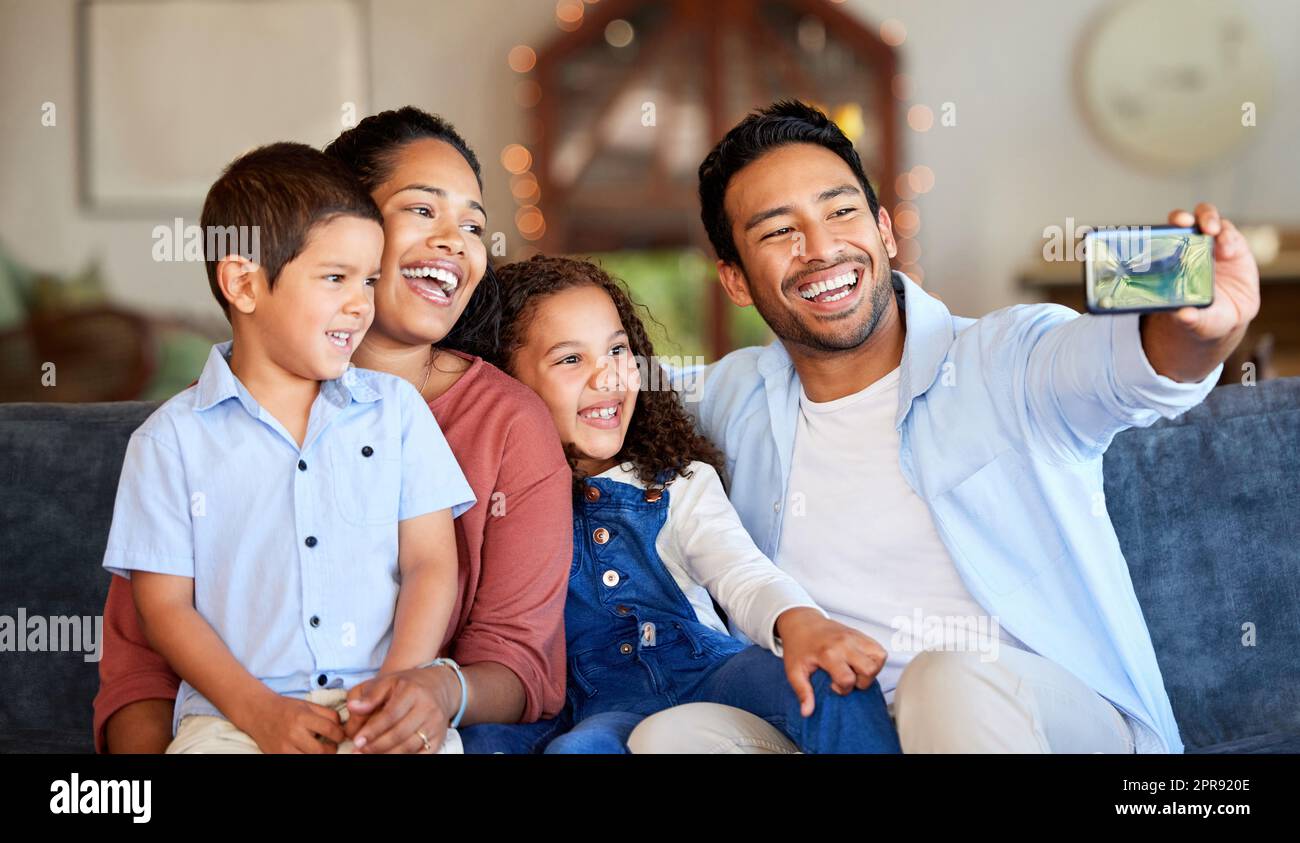 Selfie di famiglia sul divano a casa. Bellissimo giovane di etnia indiana che scatta foto con la sua famiglia di razza mista sul divano del soggiorno. Uso del telefono per scattare una foto per l'album Foto Stock