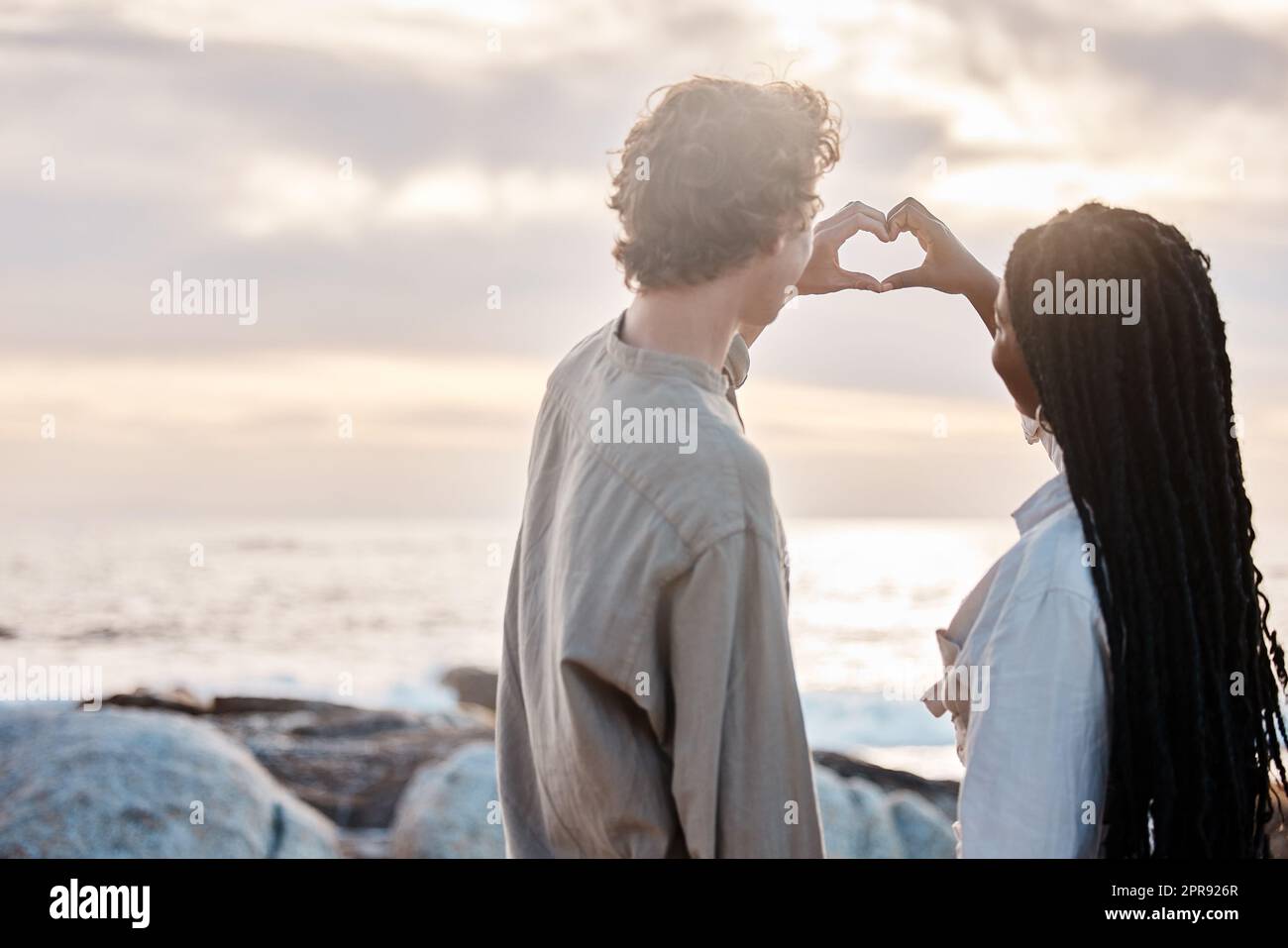 La vista posteriore di una giovane coppia multirazziale è romantica facendo un gesto di cuore con le mani alla spiaggia Foto Stock