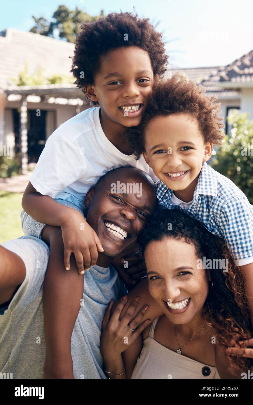 Felice famiglia afro-americana di quattro persone che si diverte prendendo selfie al sole. Genitori spensierati che trasportano figli per cavalcare il piggyback mentre si uniscono all'esterno. Mamma e papa' si divertono con i bambini Foto Stock