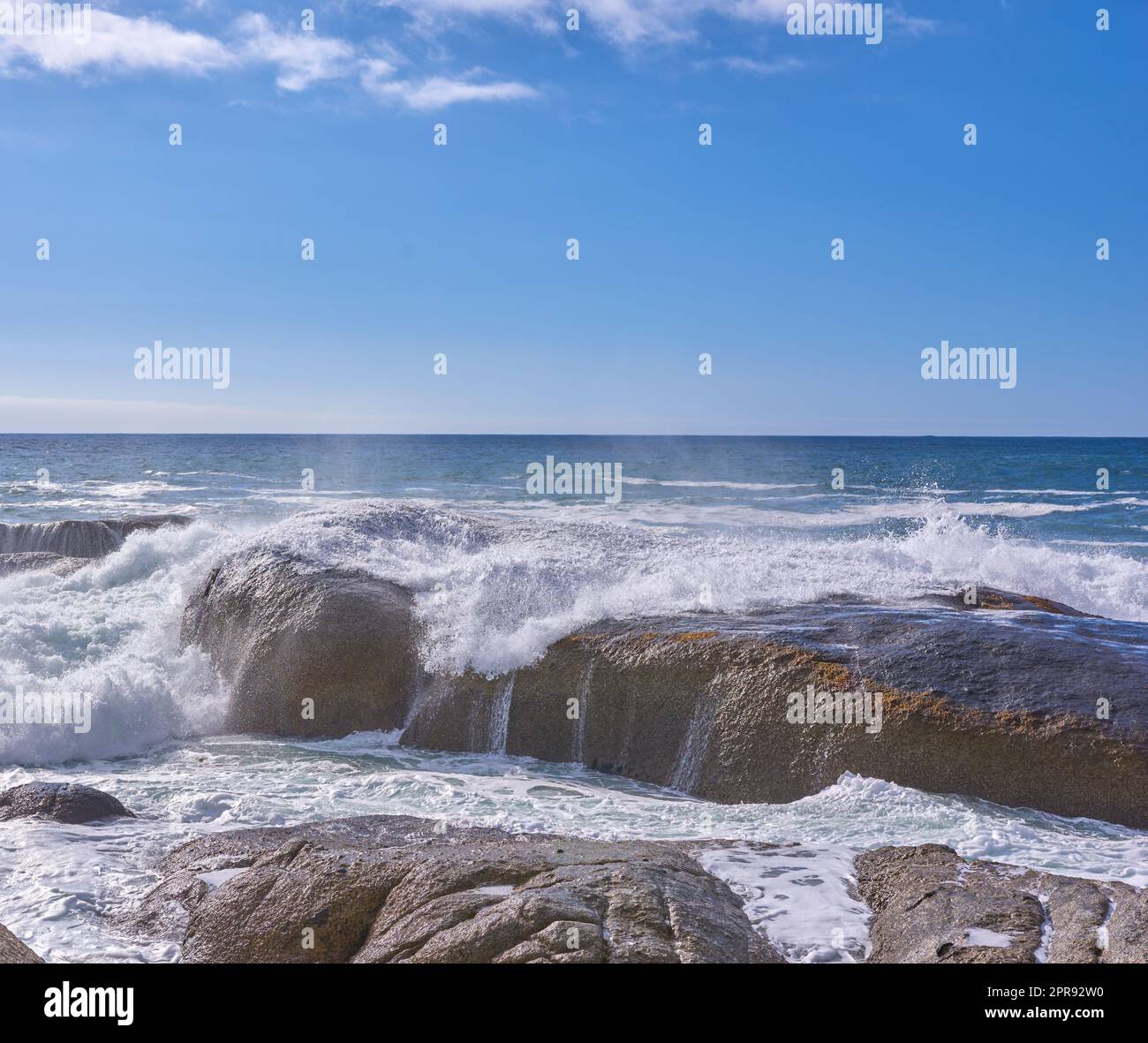 Rocce nell'oceano sotto un cielo blu nuvoloso con spazio per le copie. Paesaggio panoramico di onde sulla spiaggia che si infrangono su massi o grandi pietre nel mare in una popolare località estiva a città del Capo, in Sudafrica Foto Stock