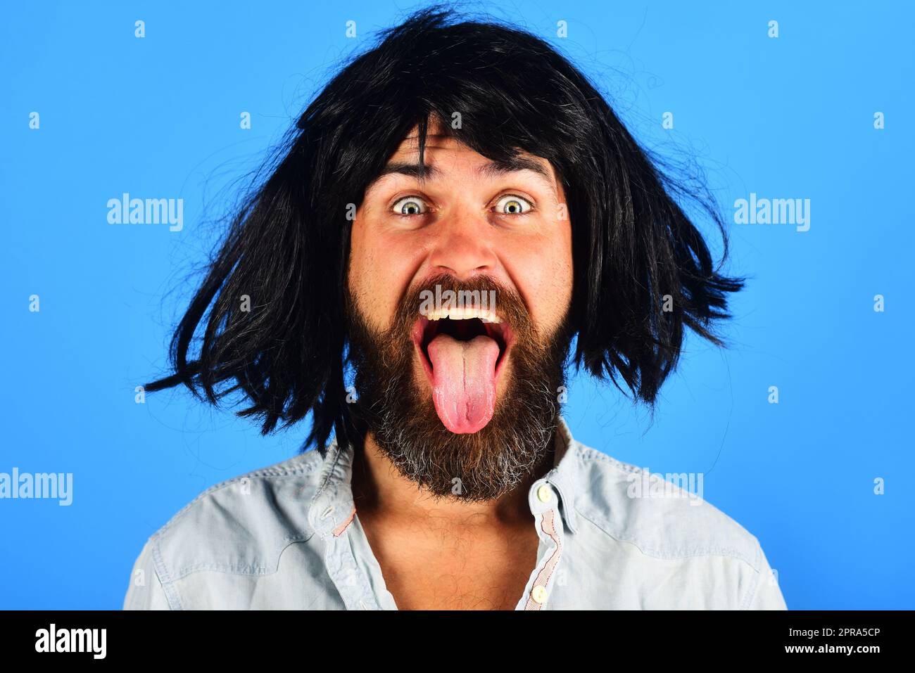 Uomo bearded divertente in parrucca nera mostra lingua. Uomo felice con barba e baffi in parrucca. Emozioni della gente. Espressione del volto. Sensazioni ed emozioni. Hipster Foto Stock