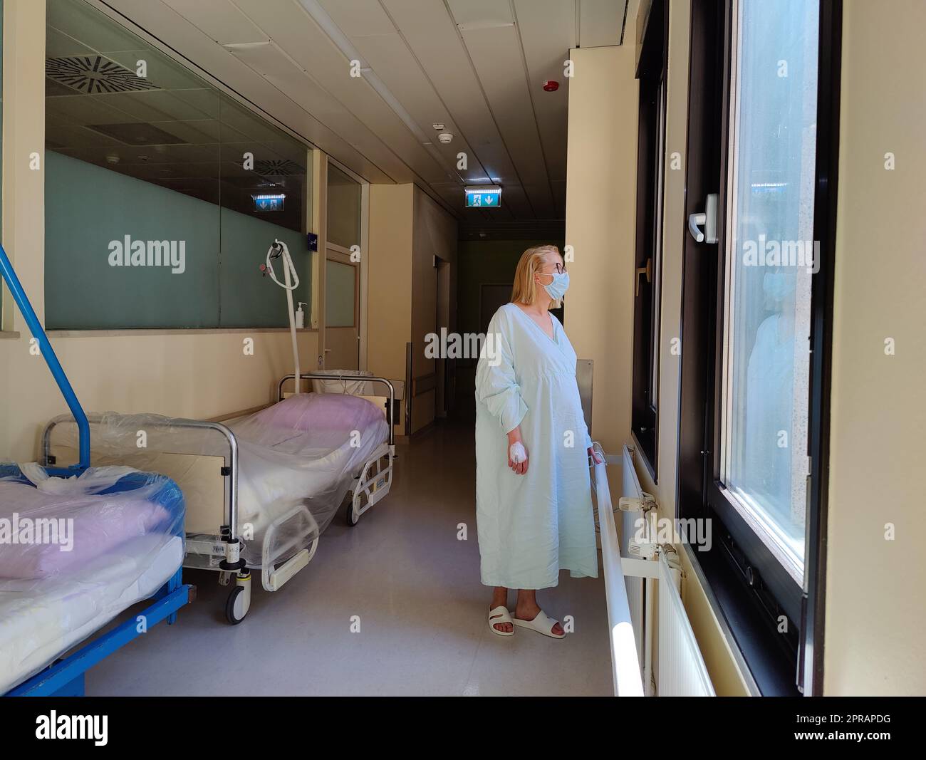 Paziente femminile solitario che indossa maschera e abito ospedaliero guardando fuori attraverso grandi finestre del corridoio dell'ospedale. Foto Stock