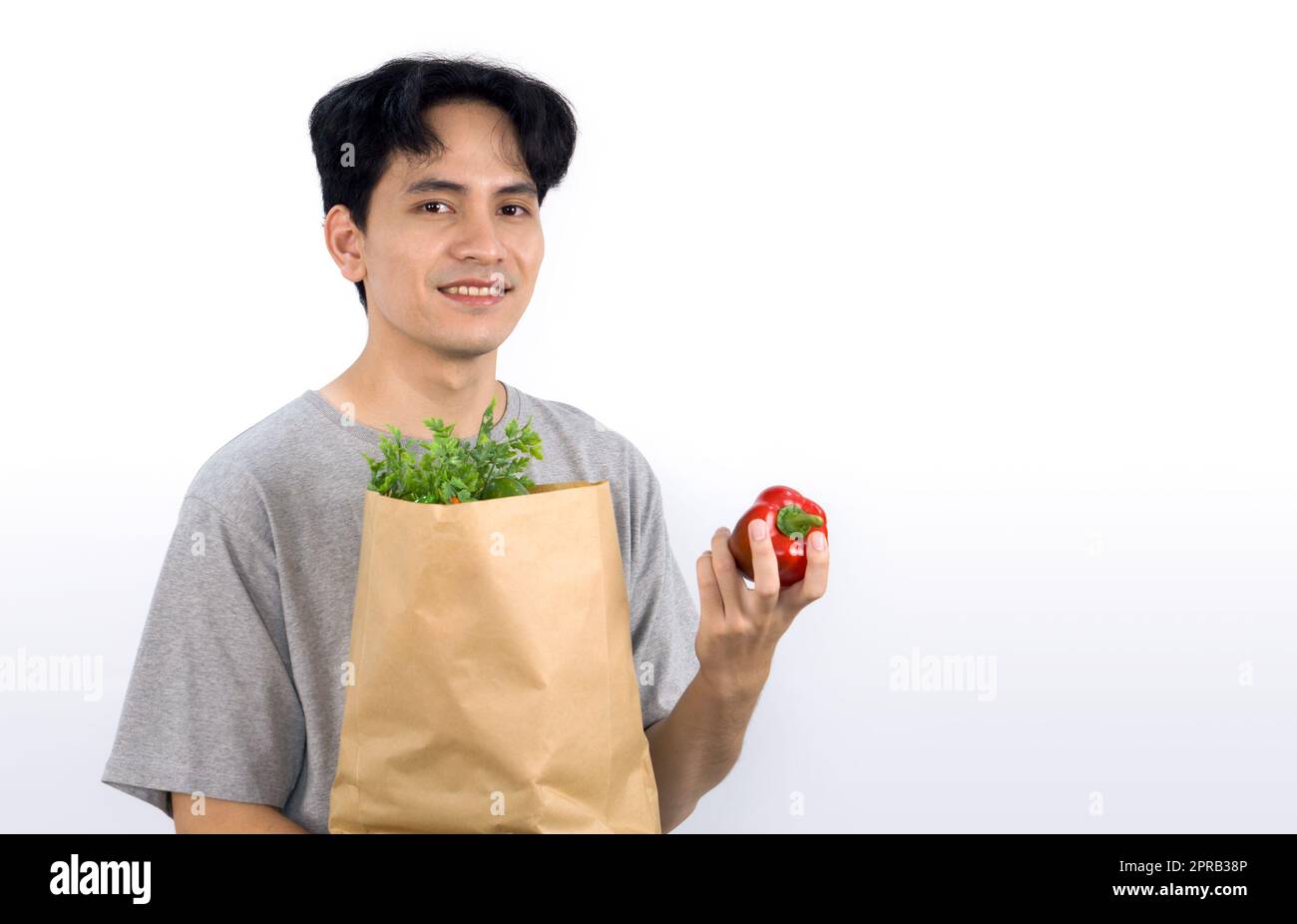 Giovane asiatico in abbigliamento casual, sorridi, tenendo in mano il peperone rosso e portando con sé un sacchetto di carta pieno di verdure. Ritratto su sfondo bianco con luce da studio. Foto Stock