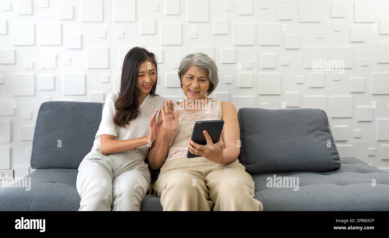 La giovane donna asiatica e la nonna agitavano la mano attraverso la comunicazione remota attraverso un computer tablet nel soggiorno. Famiglia felice che vive insieme Foto Stock