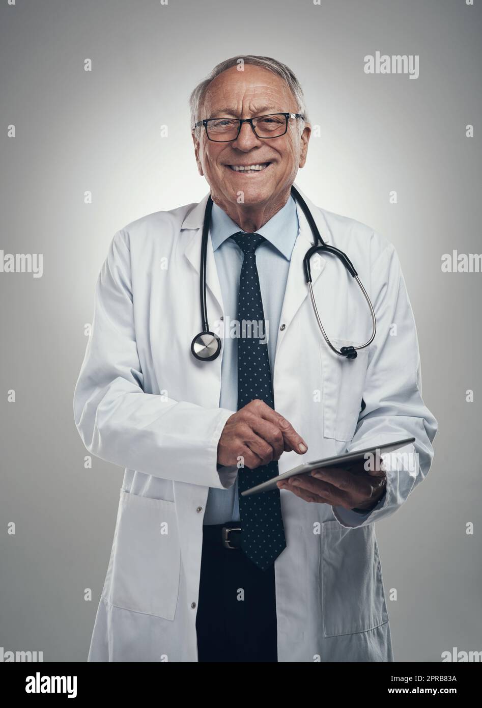 Mi piace vedere un sorriso sui volti dei miei pazienti. Un medico anziano di sesso maschile che usa un tablet digitale in uno studio su sfondo grigio. Foto Stock