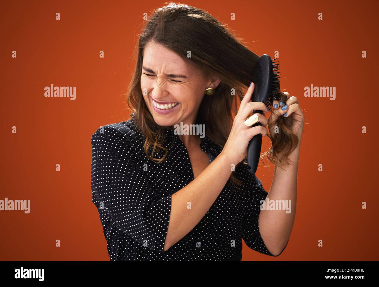 Come ha ottenuto questo nodty. Una giovane donna attraente che si alza su uno sfondo rosso nello studio e che lotta per spazzolarle i capelli. Foto Stock