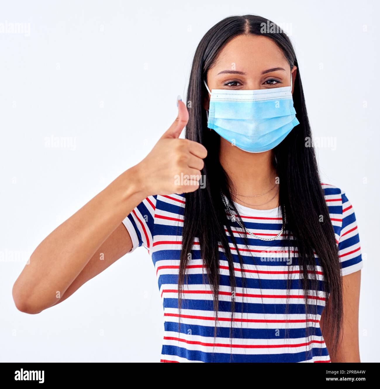 Mantenere una mentalità positiva nella pandemia. Ritratto di studio di una giovane donna che indossa una maschera facciale e mostra i pollici su uno sfondo bianco. Foto Stock