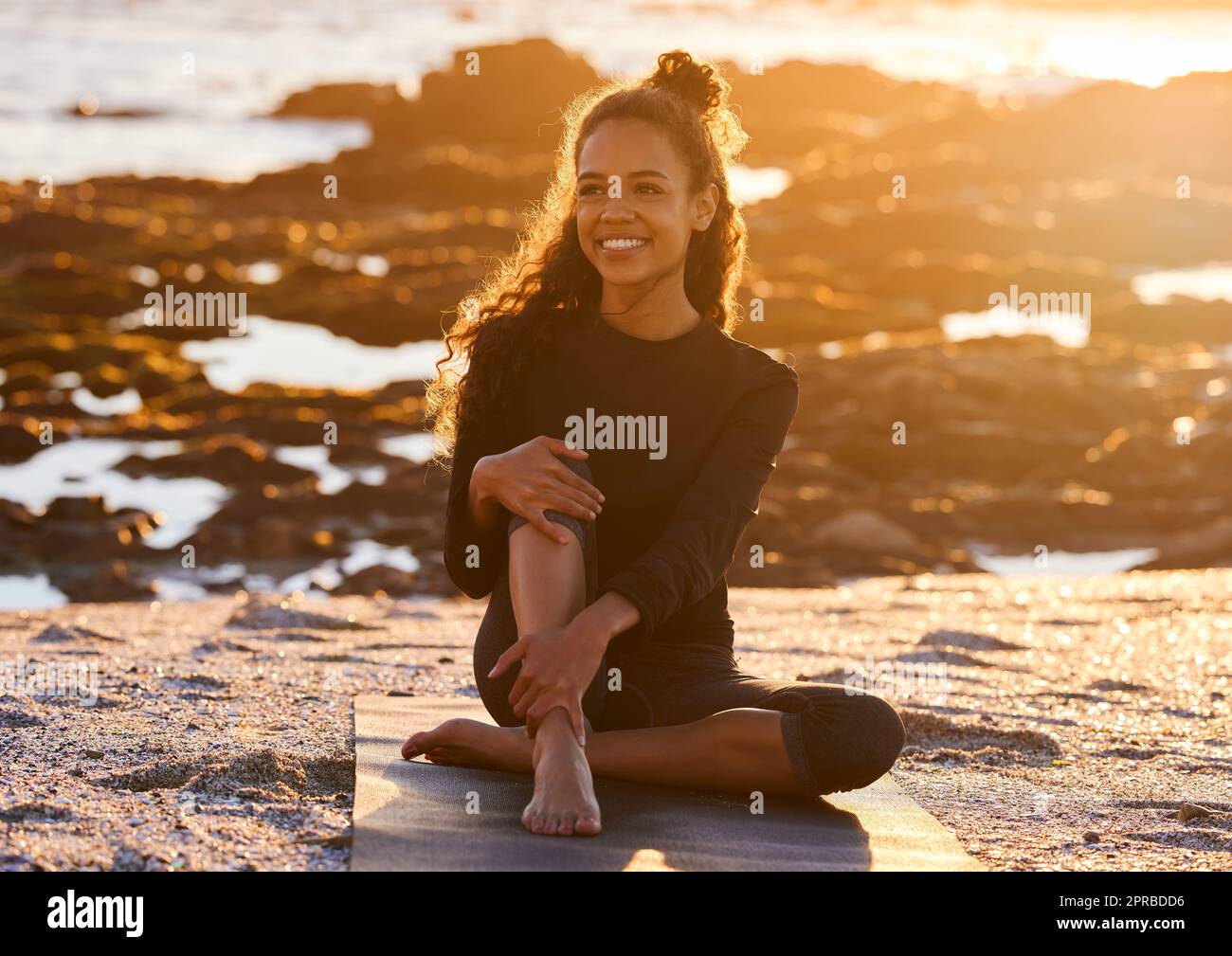 Lo yoga è la pratica di essere presente in ogni momento. Una giovane donna attraente seduta sul suo tappeto yoga durante una sessione di yoga sulla spiaggia al tramonto. Foto Stock