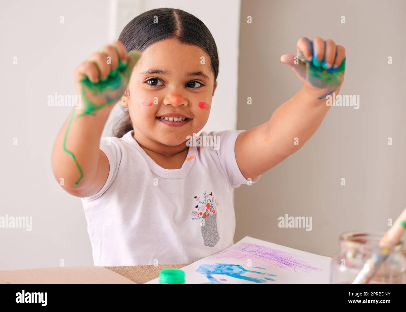 Più si presenta, più è divertente. Una bambina adorabile seduta a casa con vernice ad acqua macchiata sulle mani. Foto Stock