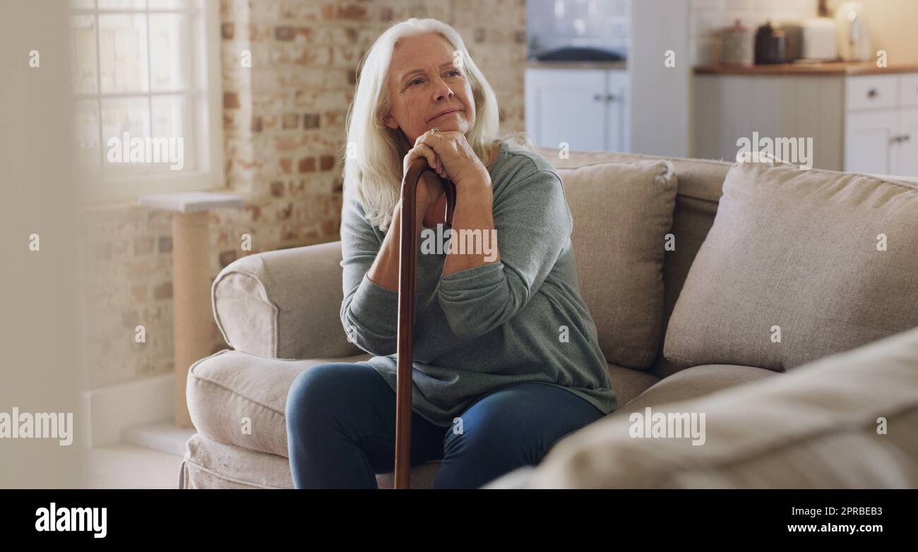 Ho tutto il tempo di pensare al mondo: Una donna anziana seduta da sola sul divano a casa e che guarda contemplativa mentre tiene il bastone da passeggio. Foto Stock