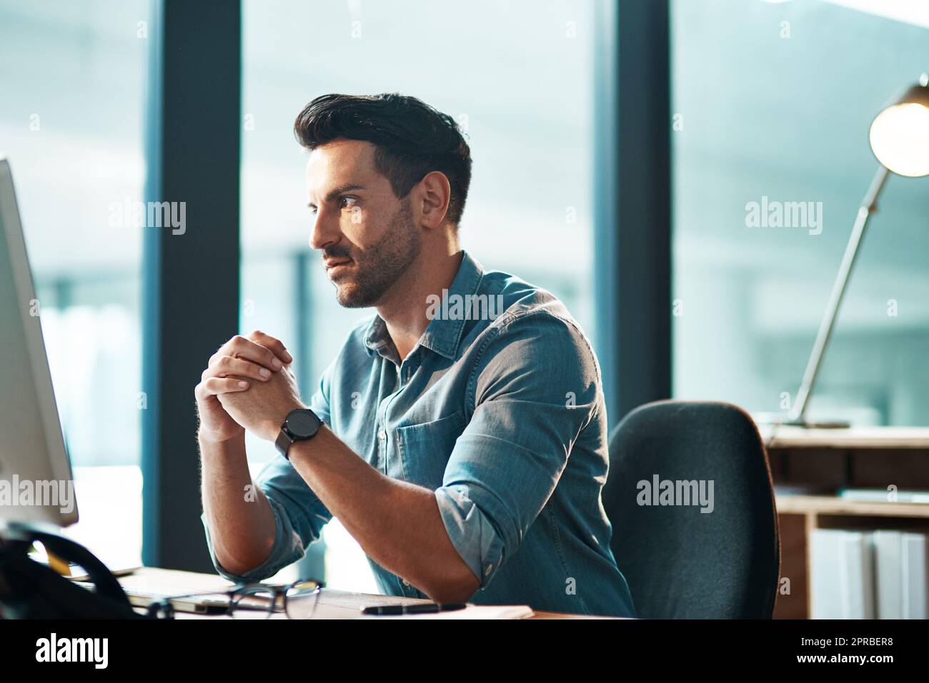 Uomo d'affari serio, impegnato e pensante che lavora da solo alla scrivania del computer all'interno di un moderno ufficio vicino a una finestra. Imprenditore, manager o capo preoccupato che leggesse e-mail e sembrasse stressato Foto Stock