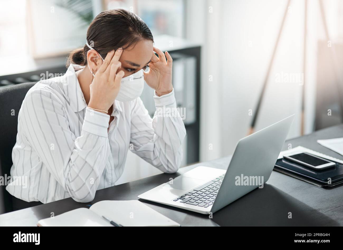 Tempi instabili portano ad un aumento dello stress: Una giovane donna d'affari mascherata che guarda stressata mentre lavora alla sua scrivania in un ufficio moderno. Foto Stock