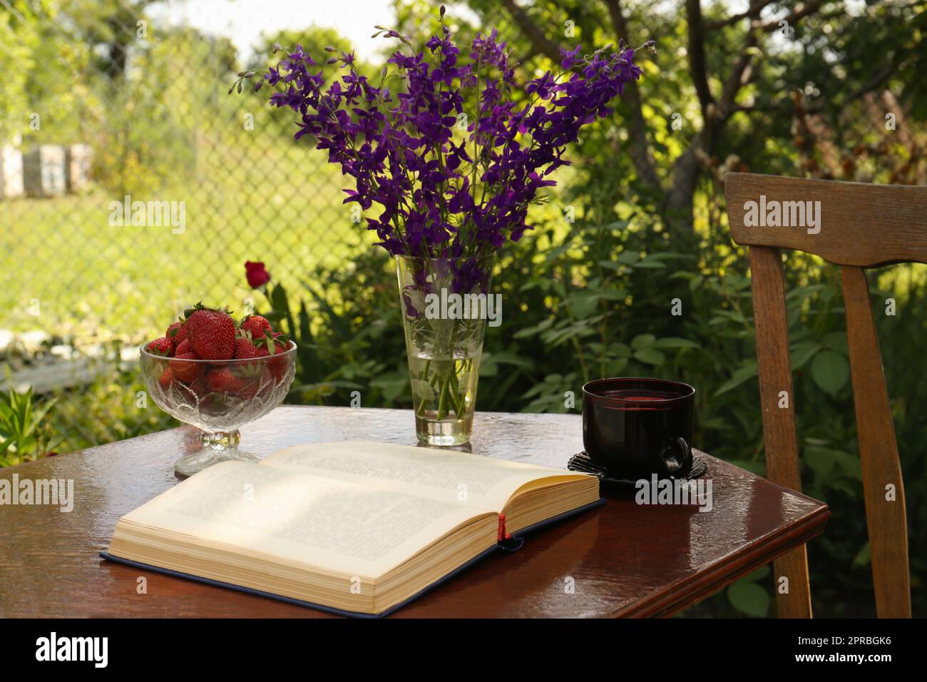 Libro aperto, tazza di tè, fragole e bellissimi fiori selvatici sul tavolo in giardino Foto Stock