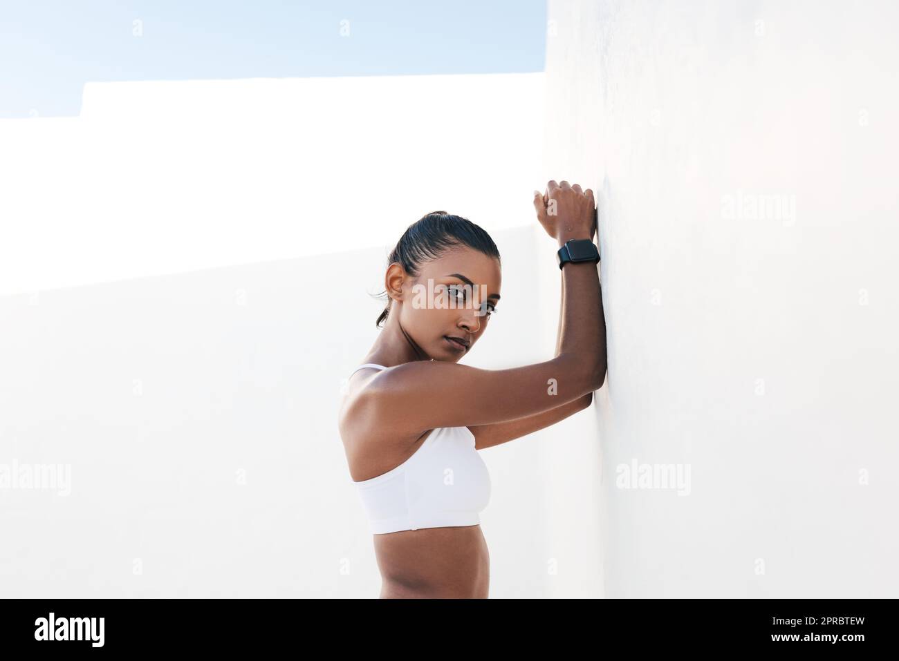 Ritratto di una parete bianca inclinata femmina adatta e guardando direttamente una fotocamera. Influenzatore di fitness che fa una breve pausa durante un allenamento. Foto Stock