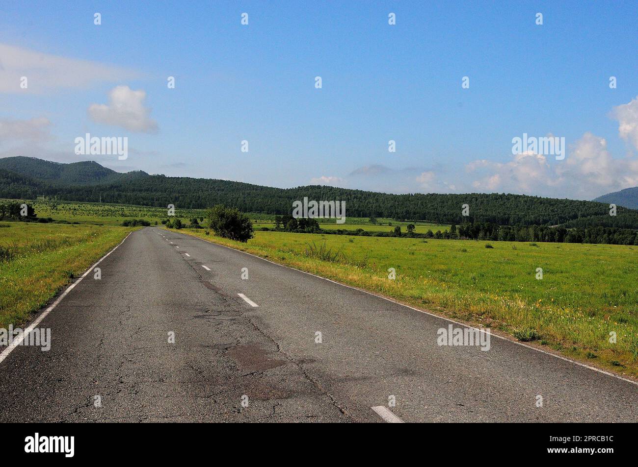 Una strada asfaltata a due corsie con crepe passa attraverso una valle pianeggiante con cespugli sparsi verso alte colline in una giornata di sole estate. Khakassia, Siberia, Russia. Foto Stock