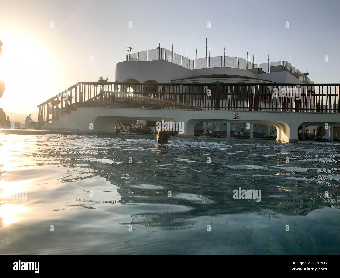 Un grande edificio in pietra con un ponte pedonale con archi sopra la piscina vicino all'acqua, una piscina contro un cielo blu e un grande sole, un tramonto nel ev Foto Stock