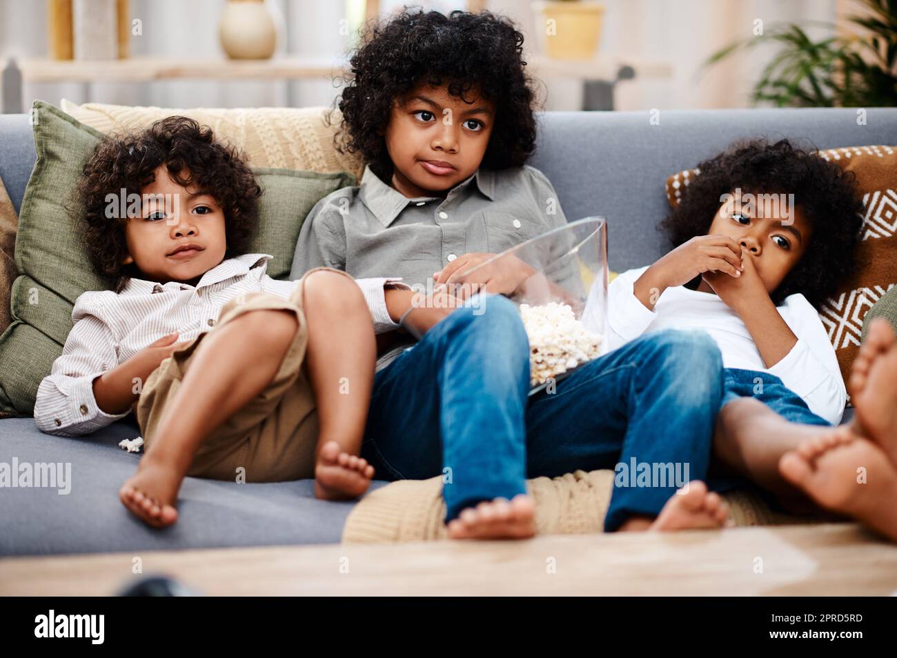 Ora cartoon. Scatto completo di tre adorabili ragazzi che mangiano popcorn e guardano la tv insieme a casa. Foto Stock