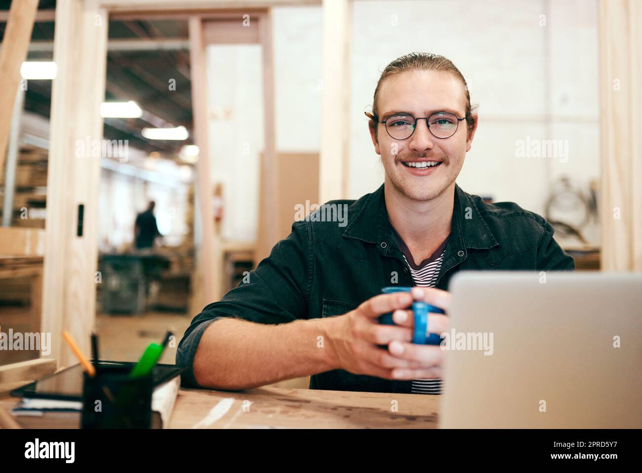 Una tazza di caffè calda fa andare la giornata. Ritratto di un bel carpentiere bere caffè e lavorare su un computer portatile all'interno della sua officina. Foto Stock