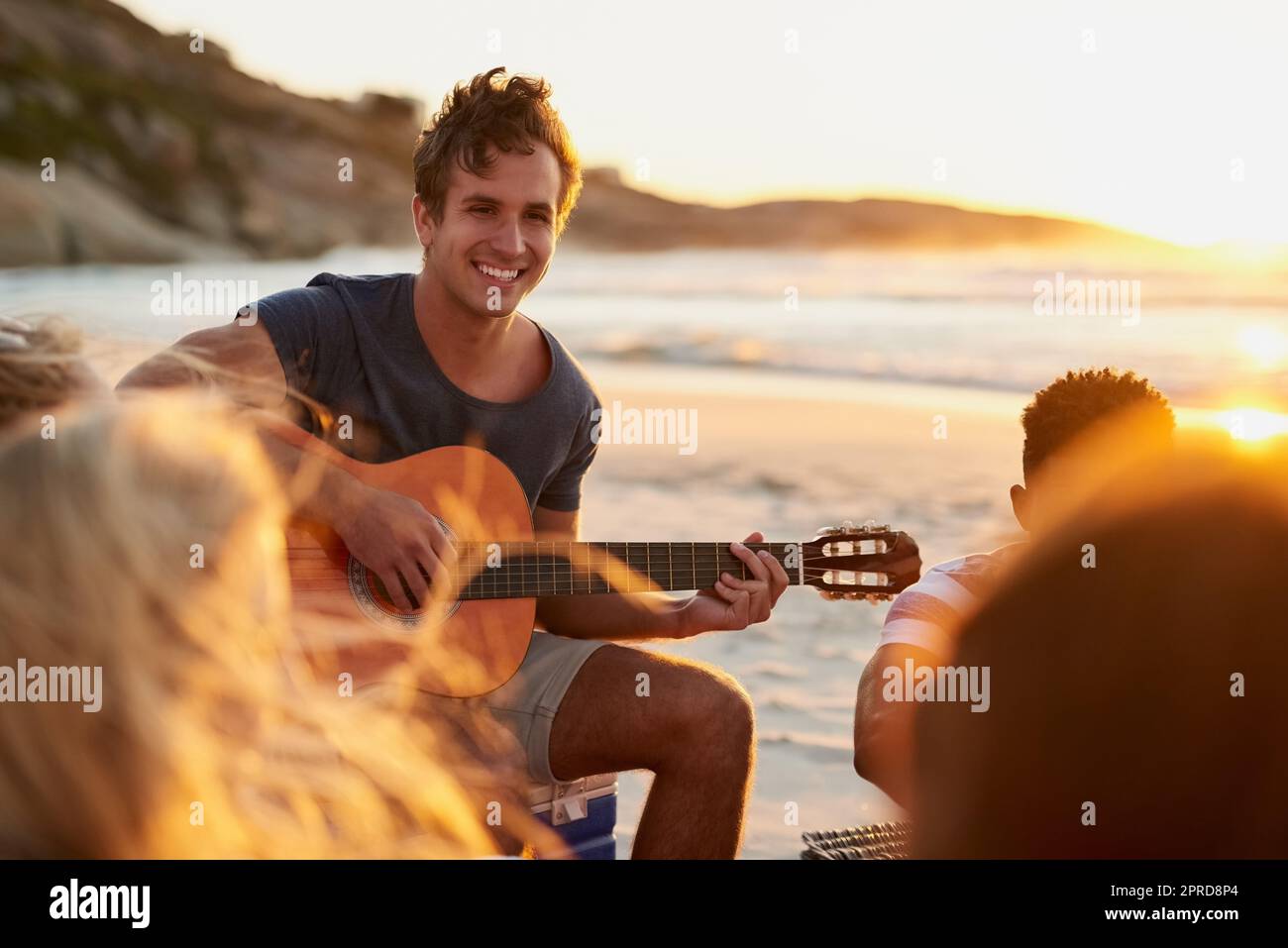 La musica e l'oceano sono davvero tutto ciò di cui abbiamo bisogno: Un uomo che suona la chitarra mentre si siede in spiaggia con i suoi amici. Foto Stock