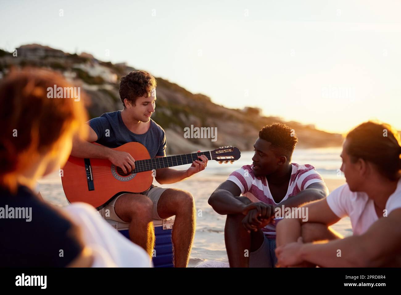 La musica è ciò che unisce le persone: Un uomo che suona la chitarra mentre si siede in spiaggia con i suoi amici. Foto Stock