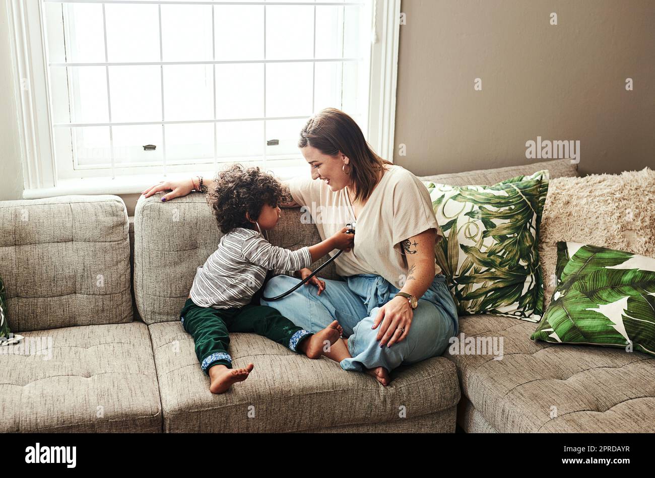 Abbiamo un medico in preparazione. una giovane donna allegra e suo figlio che giocano con uno stetoscopio mentre sono seduti su un divano a casa durante il giorno. Foto Stock