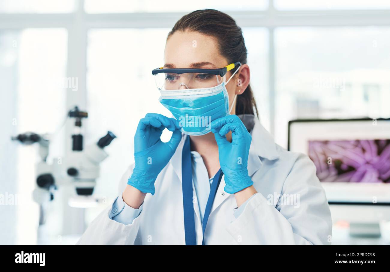 Assicurandosi che il relativo correttamente. Una scienziata giovane e irriconoscibile che mette su una maschera protettiva del viso all'interno di un laboratorio durante il giorno. Foto Stock