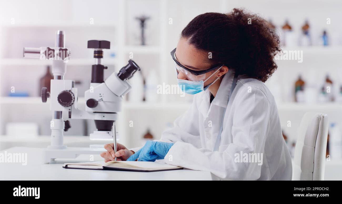 Un giovane scienziato che scrive appunti mentre usa un microscopio in laboratorio. Foto Stock