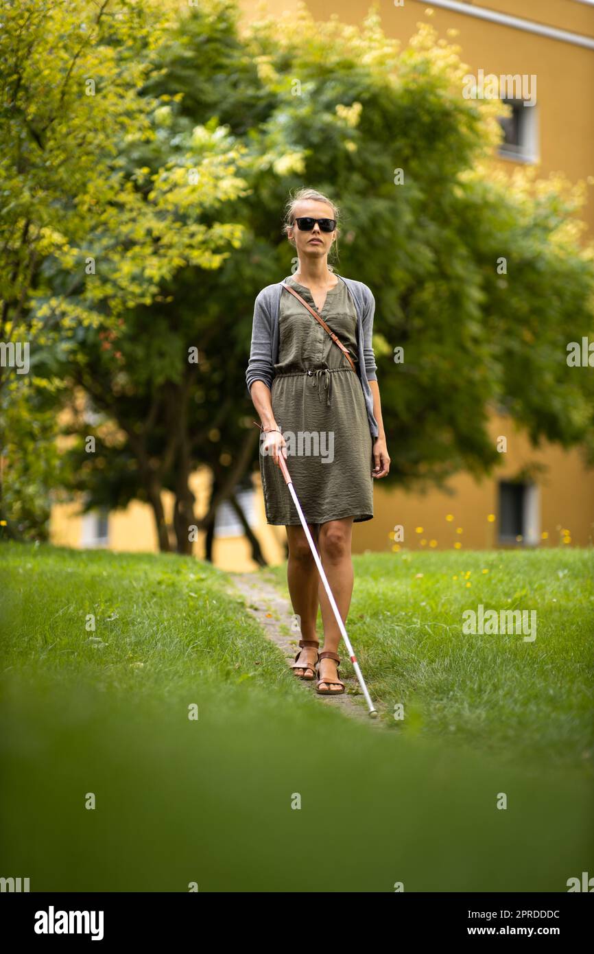 Donna cieca che cammina per le strade della città, utilizzando la sua canna bianca per navigare meglio lo spazio urbano e per raggiungere la sua destinazione in modo sicuro Foto Stock