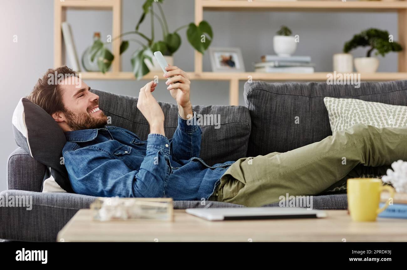 Sabato è per il divano e ricordi divertenti. Un bel giovane uomo che usa uno smartphone mentre si sdraia sul divano a casa. Foto Stock