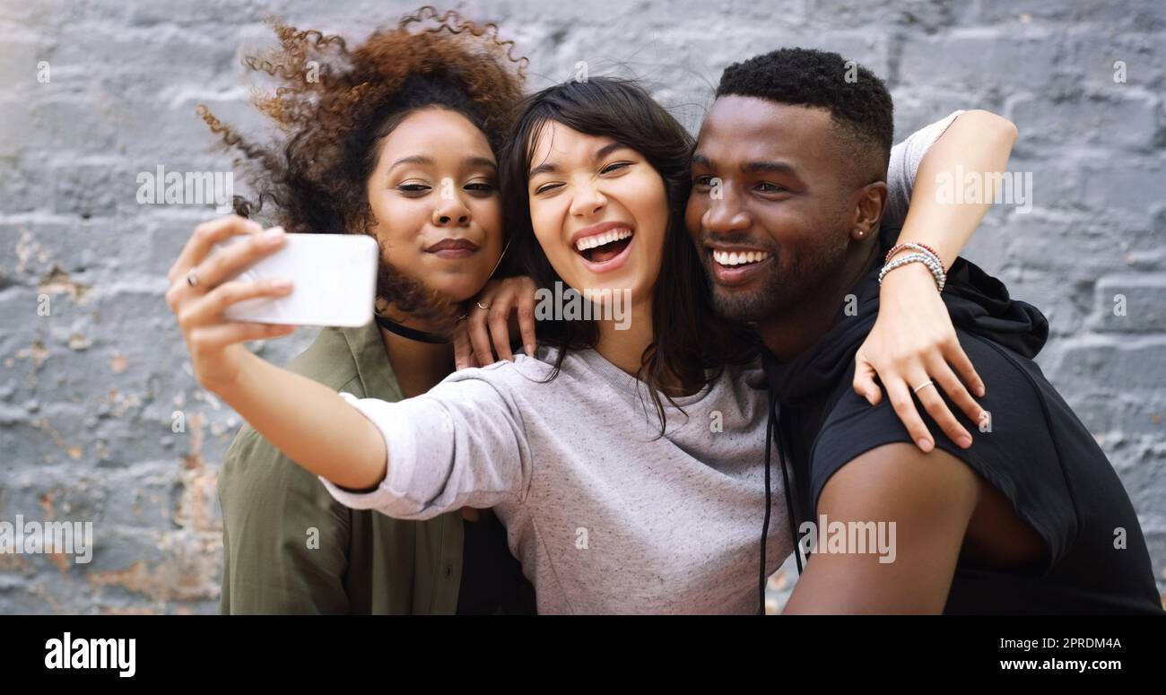 I suoi selfie tempo ragazzi, colpire una posa. Un gruppo di amici prendere un selfie insieme mentre in posa all'aperto contro una parete di mattoni grigi. Foto Stock