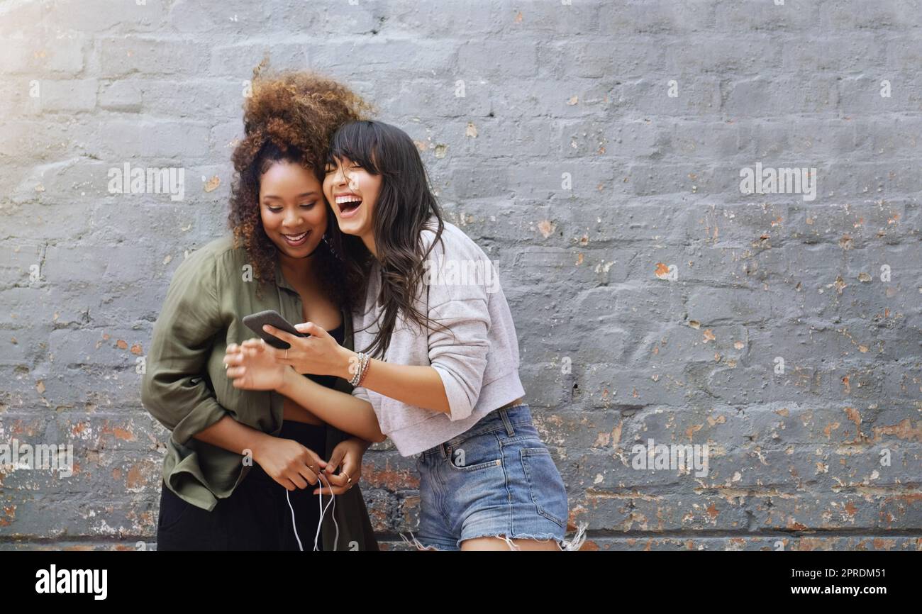 I social media ci fanno sempre ridere ad alta voce. Due giovani donne allegre che usano un cellulare insieme mentre si posano all'aperto contro un muro di mattoni grigi. Foto Stock