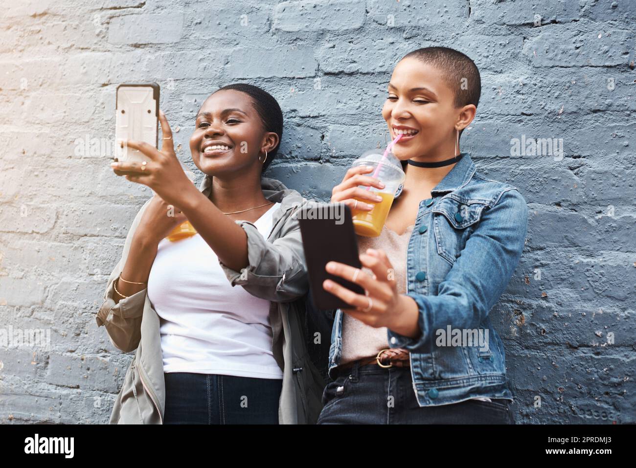 Il sovraccarico di cuteness. Due giovani donne in piedi accanto ad un edificio sorridendo e prendendo selfie mentre tengono le loro bevande fredde. Foto Stock