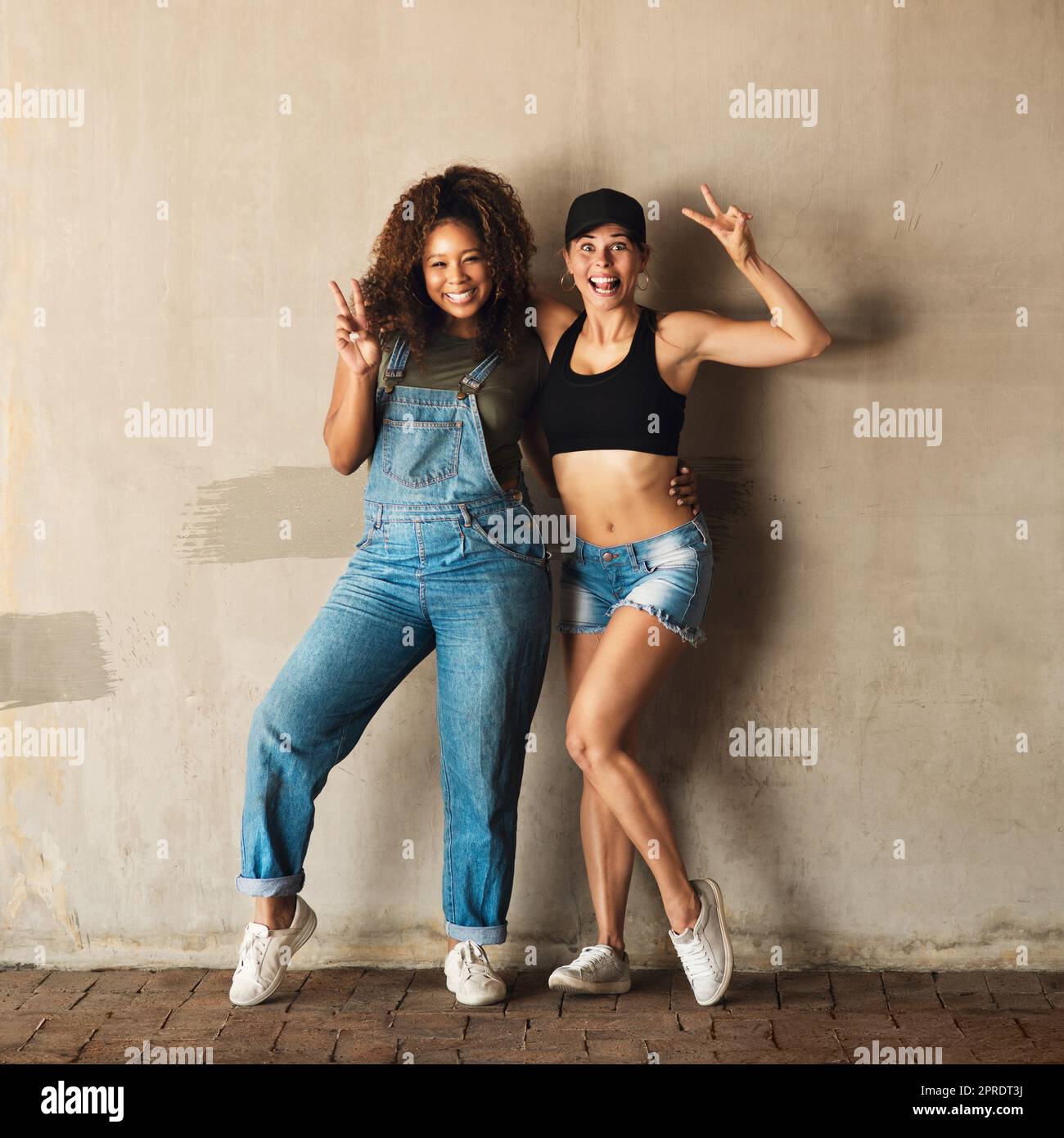 Veniamo in pace. Ritratto di due giovani donne allegre in posa per una foto mentre si appoggiano a una parete all'esterno durante il giorno. Foto Stock